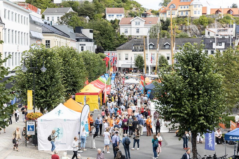 Mennesker som myldrer i en travel gate mellom standboder i en sommerby. Løvtrær og trehus. Foto.
