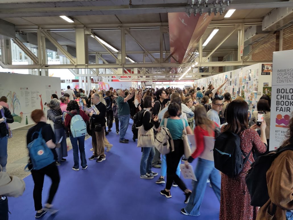 myldreområdepå bokmesse, folk som går i alle retninger med bærenett og sekker på rygg og skuldre, noen fotograferer plakater på vegger. 