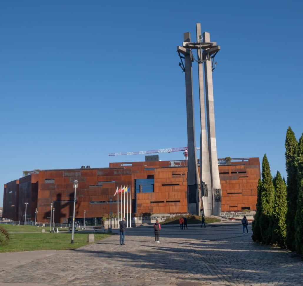 Åpen plass foran større bygg med betongmonument i forgrunnen, spredt tilkomst av mennesker i bildet. Foto.