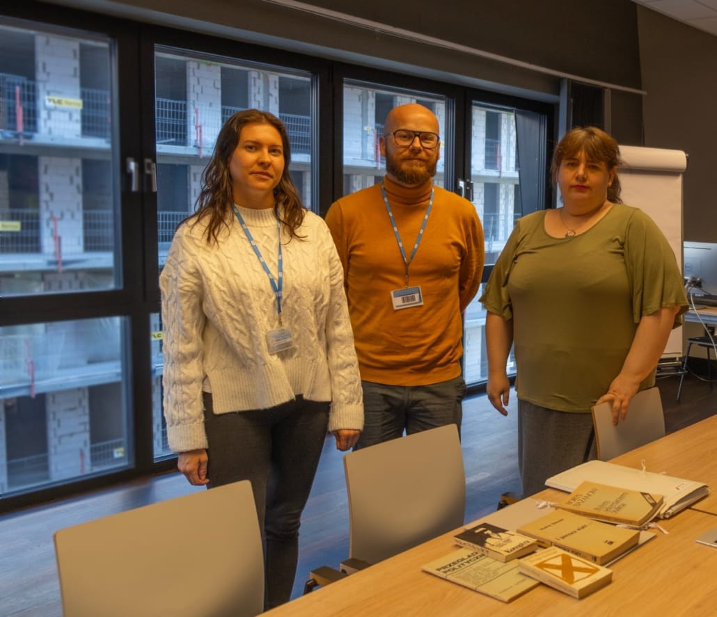 Tre personer/ kolleger kvinne-mann-kvinne- ved biblioteket fotografert sammen forfra, kontoromgivelser, står foran et bord med bøker. Foto.