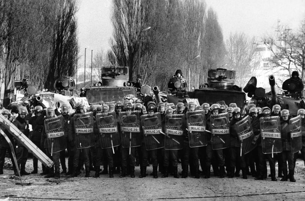 Bilde i svart/hvitt av polititropp med hjelmer og skjold, bak dem tanks og større militærkjøretøy, de danner en sperring/front. Foto.