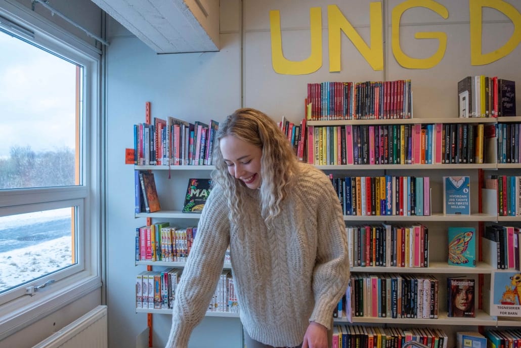 kvinne ved bord stående foran bokhylle og bokstavene UNG på veggen bak henne. Vindu. Foto. 